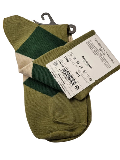 A pair of knit socks with a green and beige argyle pattern, shown with a white paper tag attached. The socks are photographed on a white sheet of paper with a wooden surface visible in the top-left corner.