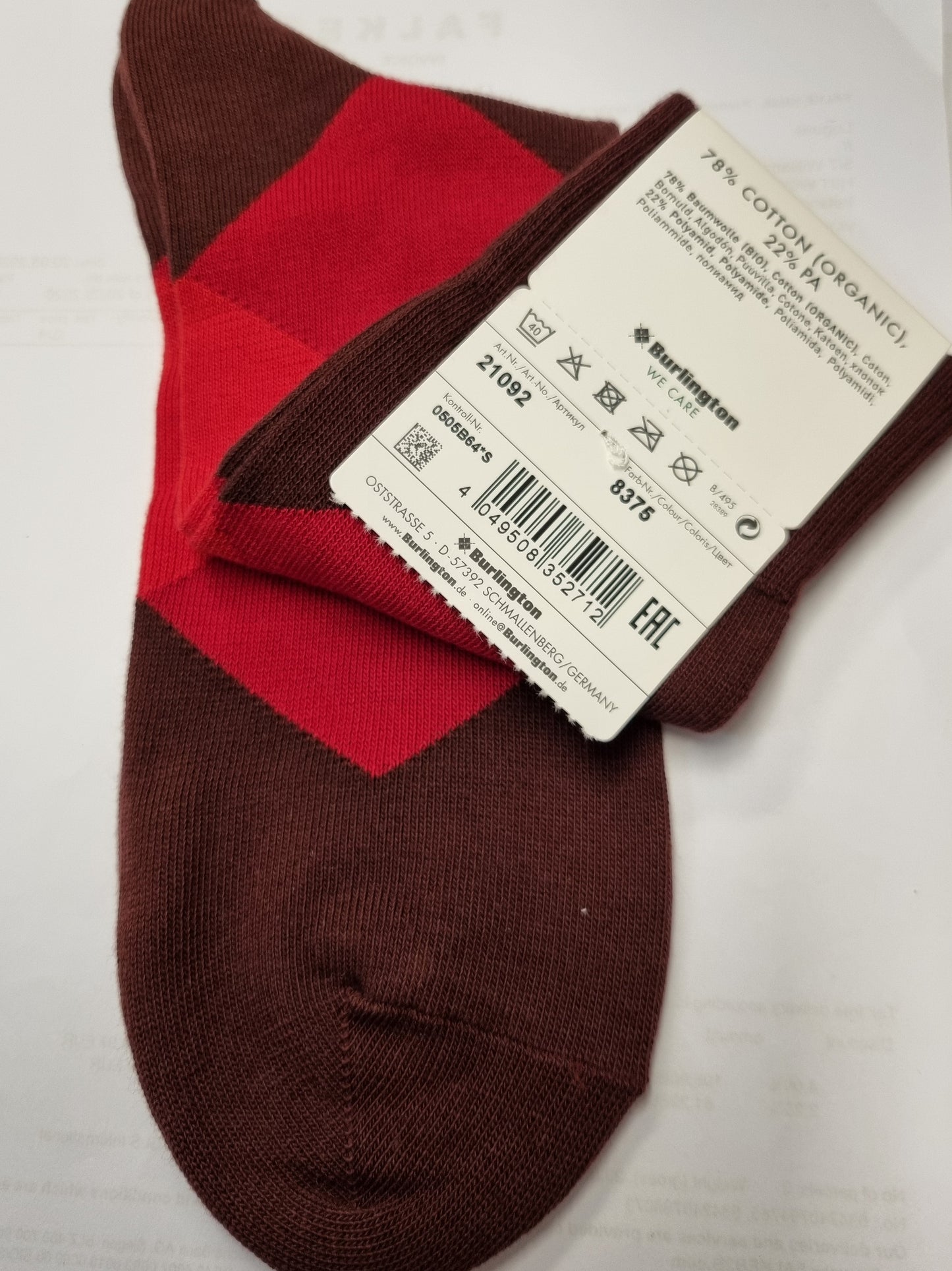 A pair of knit socks with a dark brown base and a red chevron pattern, featuring a white product tag attached. The socks are laid flat on a white sheet of paper.